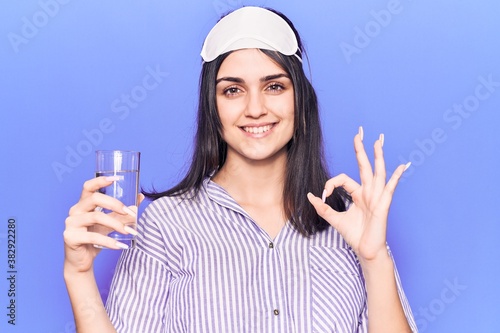 Young beautiful girl wearing sleep mask drinking glass of water doing ok sign with fingers, smiling friendly gesturing excellent symbol