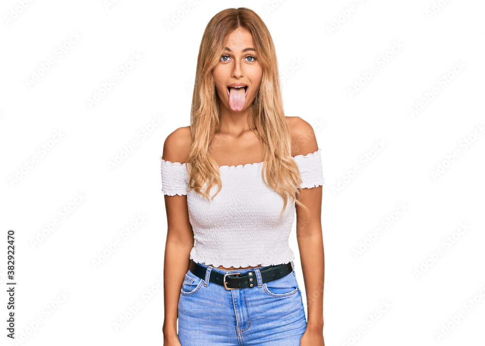 Beautiful blonde young woman wearing casual white tshirt sticking tongue out happy with funny expression. emotion concept. Beautiful blonde young woman wearing casual white tshirt sticking tongue out happy with funny expression. emotion concept.