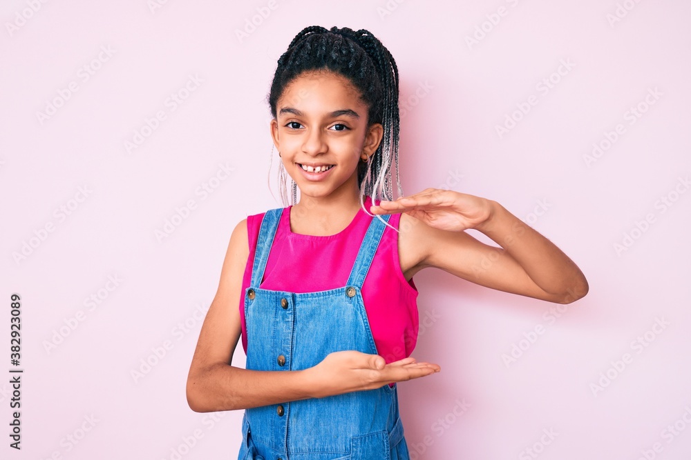 Young african american girl child with braids wearing casual clothes over pink background gesturing with hands showing big and large size sign, measure symbol. smiling looking at the camera. Young african american girl child with braids wearing casual clothes over pink background gesturing with hands showing big and large size sign, measure symbol. smiling looking at the camera.