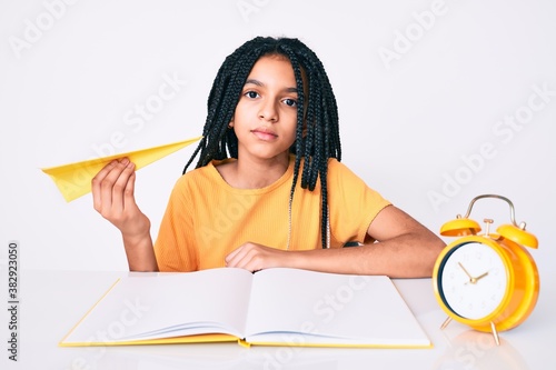 Young african american girl child with braids holding paper airplane while studying thinking attitude and sober expression looking self confident photo