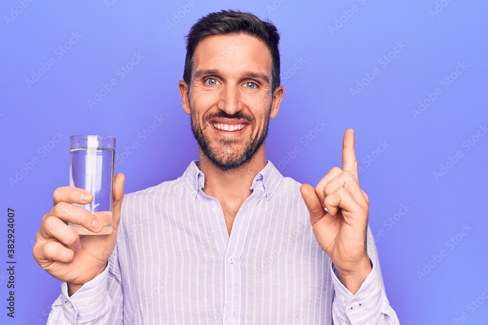 Young handsome man drinking glass of water to refreshment over isolated purple background smiling happy pointing with hand and finger to the side Young handsome man drinking glass of water to refreshment over isolated purple background smiling happy pointing with hand and finger to the side