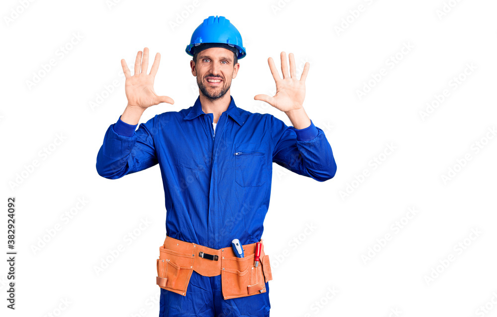 Young handsome man wearing worker uniform and hardhat showing and pointing up with fingers number ten while smiling confident and happy.