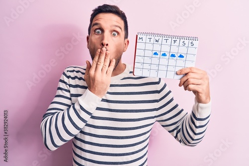 Young handsome man holding weather calendar showing rainy week over pink background covering mouth with hand, shocked and afraid for mistake. Surprised expression photo