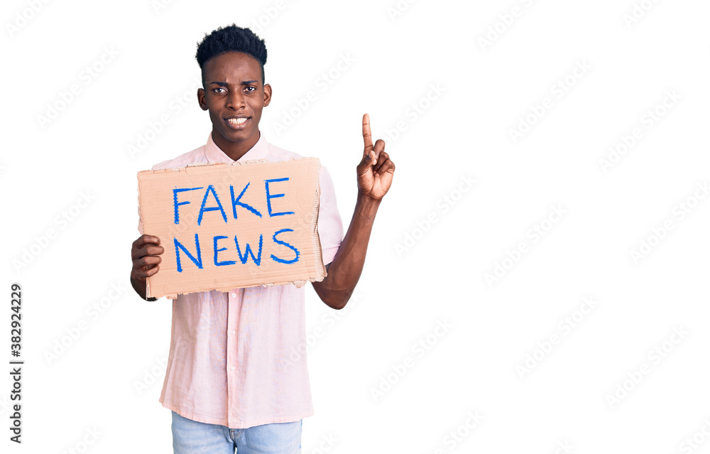 Young african american man holding fake news banner surprised with an idea or question pointing finger with happy face, number one Young african american man holding fake news banner surprised with an idea or question pointing finger with happy face, number one