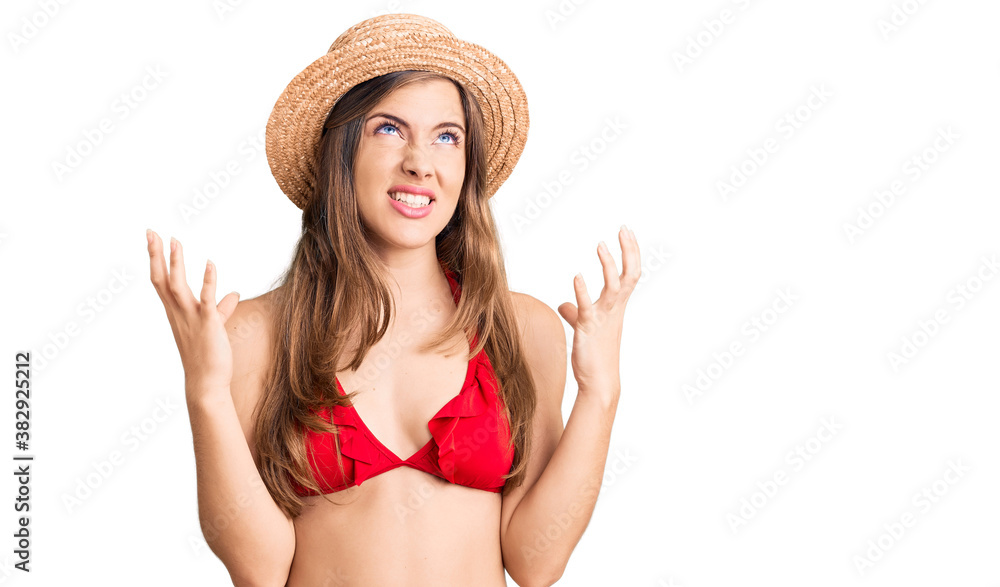 Beautiful caucasian young woman wearing bikini and summer hat crazy and mad shouting and yelling with aggressive expression and arms raised. frustration concept. Beautiful caucasian young woman wearing bikini and summer hat crazy and mad shouting and yelling with aggressive expression and arms raised. frustration concept.