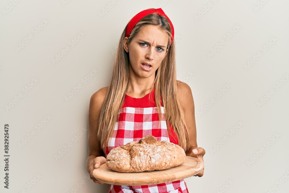 Young blonde woman wearing baker uniform holding homemade bread in shock face, looking skeptical and sarcastic, surprised with open mouth Young blonde woman wearing baker uniform holding homemade bread in shock face, looking skeptical and sarcastic, surprised with open mouth