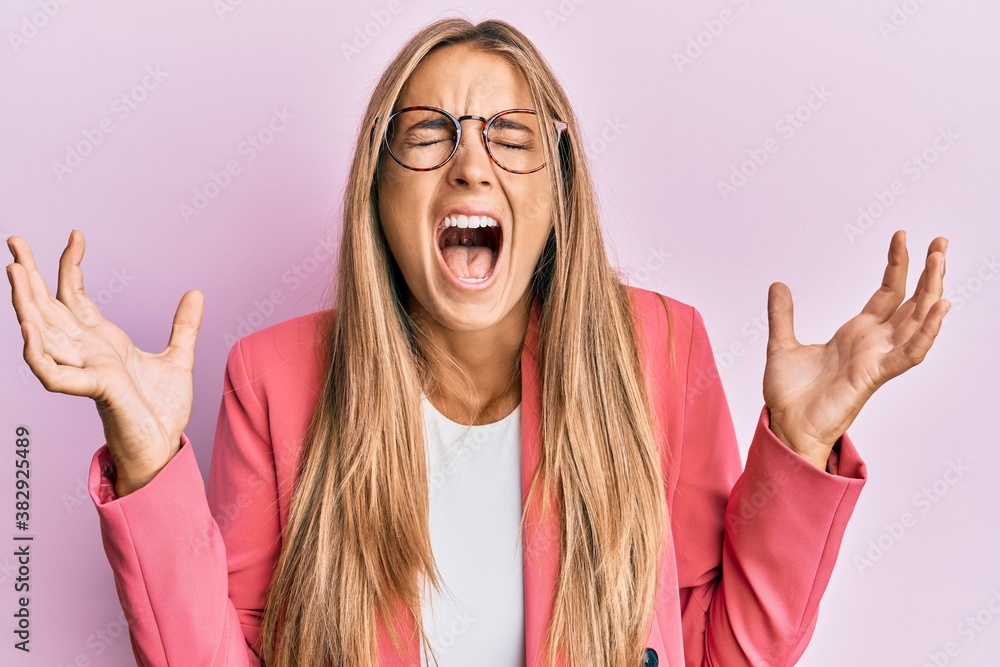 Young blonde woman wearing business style and glasses celebrating mad and crazy for success with arms raised and closed eyes screaming excited. winner concept Young blonde woman wearing business style and glasses celebrating mad and crazy for success with arms raised and closed eyes screaming excited. winner concept