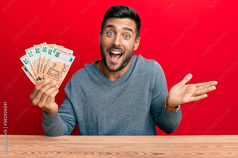 Handsome hispanic man holding bunch of 50 euro banknotes celebrating achievement with happy smile and winner expression with raised hand Handsome hispanic man holding bunch of 50 euro banknotes celebrating achievement with happy smile and winner expression with raised hand