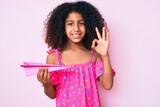 African american child with curly hair holding paper airplane doing ok sign with fingers, smiling friendly gesturing excellent symbol