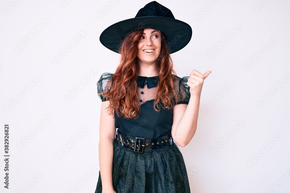 Young beautiful woman wearing witch halloween costume smiling with happy face looking and pointing to the side with thumb up. Young beautiful woman wearing witch halloween costume smiling with happy face looking and pointing to the side with thumb up.