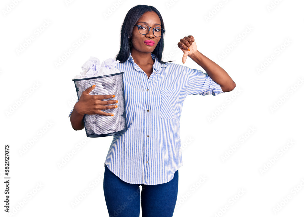 Young african american woman holding paper bin full of crumpled papers with angry face, negative sign showing dislike with thumbs down, rejection concept