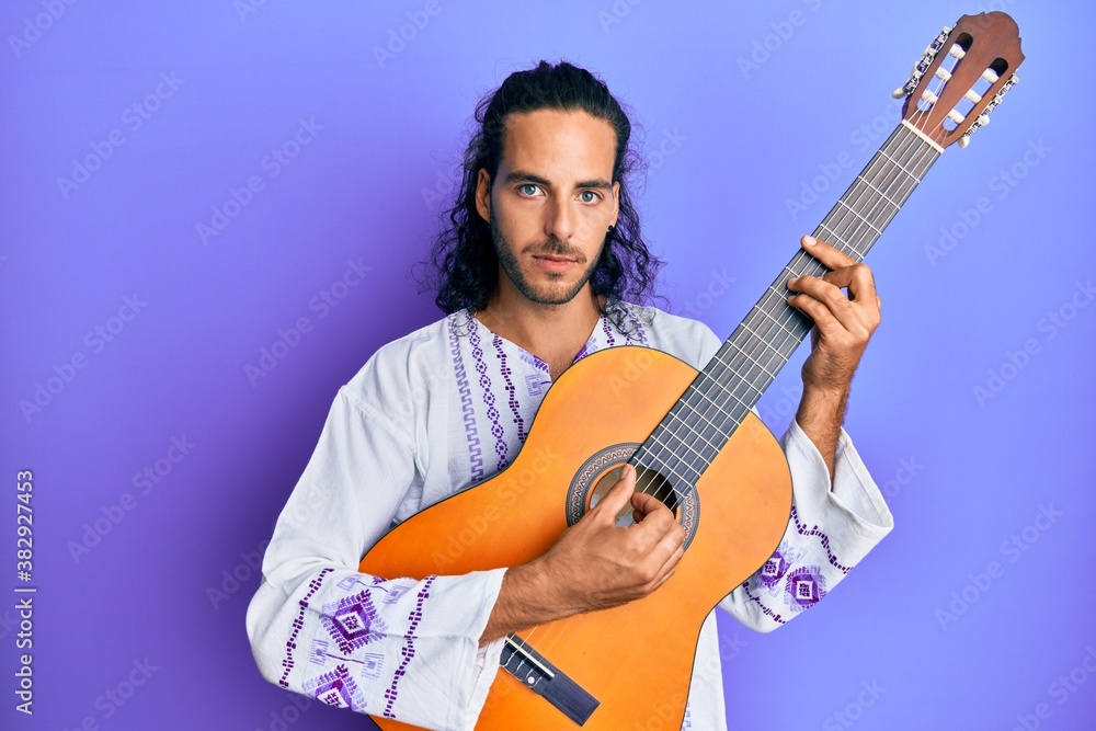 Naklejka premium Young handsome man with long hair playing classical guitar relaxed with serious expression on face. simple and natural looking at the camera.