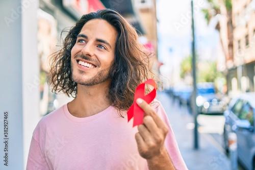 Картината върху платно Young hispanic man smiling happy holding hiv awareness red ribbon at city