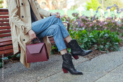 Fashionable young woman wearing beige trench coat, sweater, blue jeans and black high heel cowboy boots. She is holding burgundy colour leather handbag in hand. Street style.