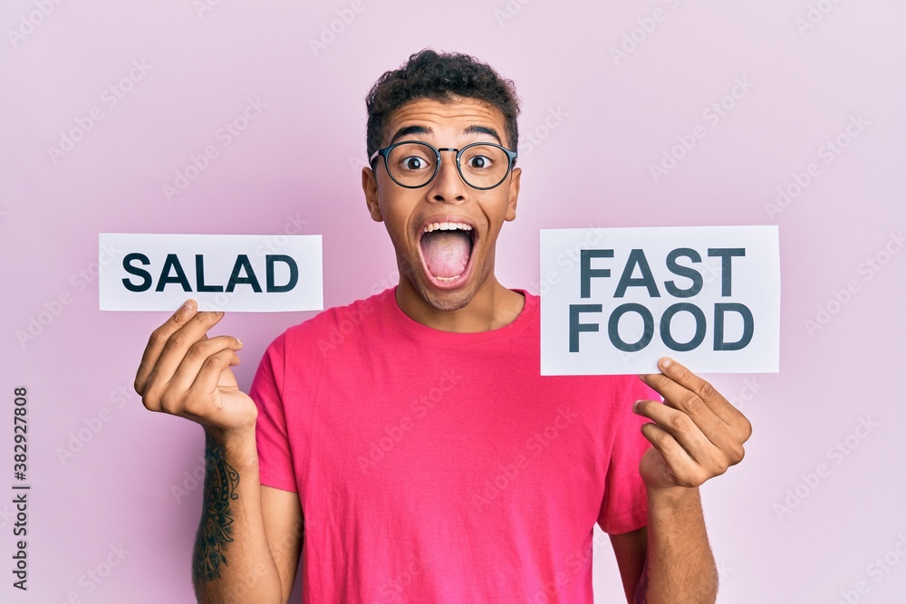 Young handsome african american man holding salad and fast food message paper celebrating crazy and amazed for success with open eyes screaming excited. Young handsome african american man holding salad and fast food message paper celebrating crazy and amazed for success with open eyes screaming excited.