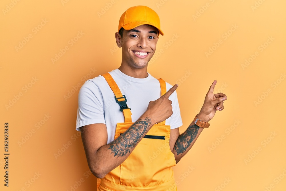 Young handsome african american man wearing handyman uniform over yellow background smiling and looking at the camera pointing with two hands and fingers to the side.