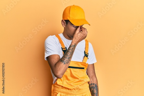 Young handsome african american man wearing handyman uniform over yellow background tired rubbing nose and eyes feeling fatigue and headache. stress and frustration concept. © Krakenimages.com