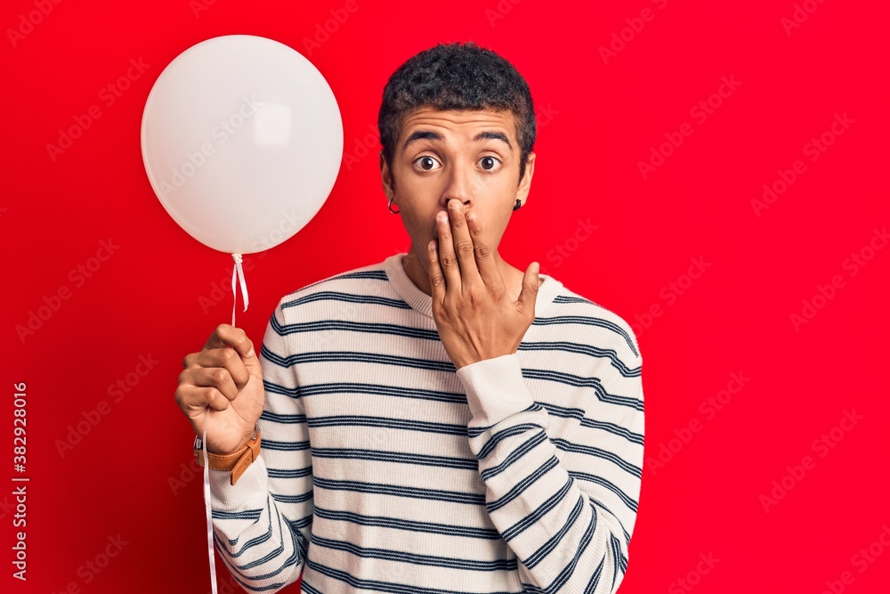 Young african amercian man holding balloon covering mouth with hand, shocked and afraid for mistake. surprised expression Young african amercian man holding balloon covering mouth with hand, shocked and afraid for mistake. surprised expression