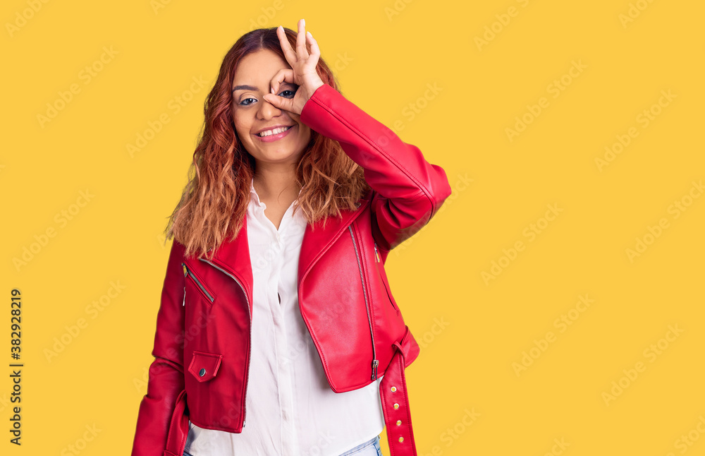 Young latin woman wearing red leather jacket smiling happy doing ok sign with hand on eye looking through fingers