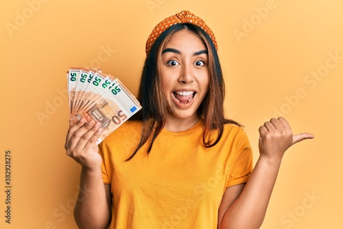 Young latin woman holding 50 euro banknotes pointing thumb up to the side smiling happy with open mouth