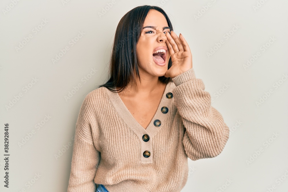 Young beautiful hispanic girl wearing casual winter sweater shouting and screaming loud to side with hand on mouth. communication concept. Young beautiful hispanic girl wearing casual winter sweater shouting and screaming loud to side with hand on mouth. communication concept.