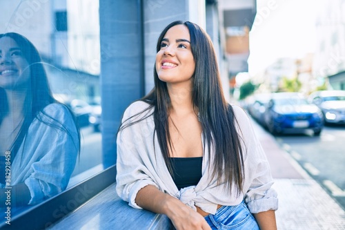 Free Download Young hispanic girl smiling happy leaning on the wall at the city. - GFXLoad
