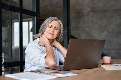 Tired stressed old mature business woman suffering from fibromyalgia neckpain working in office sitting at table. Overworked senior middle aged lady massaging neck feeling hurt pain from sedentary job photo
