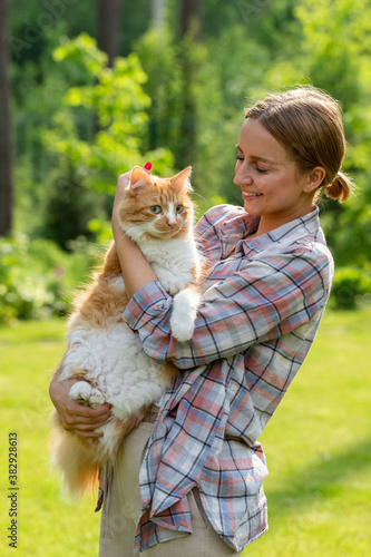 Happy woman in checked shirt hugging and embracing with tenderness and love domestic ginger cat outdoors in sunny day. Love to the animals