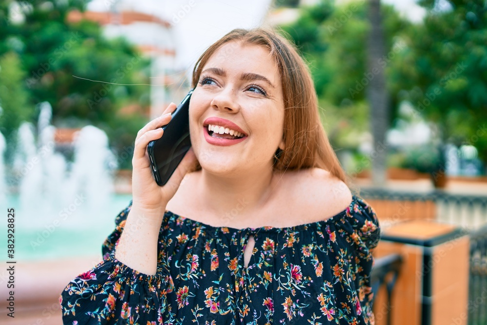 Fototapeta premium Young redhead woman talking on the smartphone walking at the park