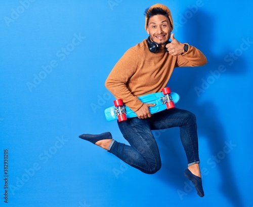 Young handsome latin man wearing wool cap jumping with smile on face. Holding skate using headphones over isolated blue background