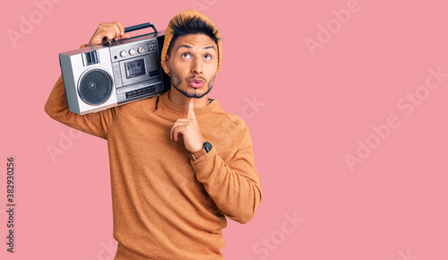 Handsome latin american young man holding boombox, listening to music thinking concentrated about doubt with finger on chin and looking up wondering