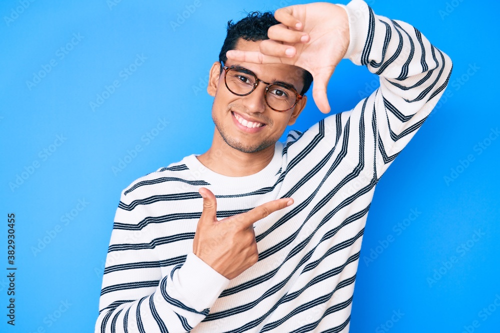 Young handsome hispanic man wearing casual sweater and glasses smiling making frame with hands and fingers with happy face. creativity and photography concept. Young handsome hispanic man wearing casual sweater and glasses smiling making frame with hands and fingers with happy face. creativity and photography concept.