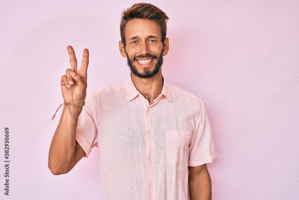 Handsome caucasian man with beard wearing casual pink shirt smiling with happy face winking at the camera doing victory sign. number two. Handsome caucasian man with beard wearing casual pink shirt smiling with happy face winking at the camera doing victory sign. number two.