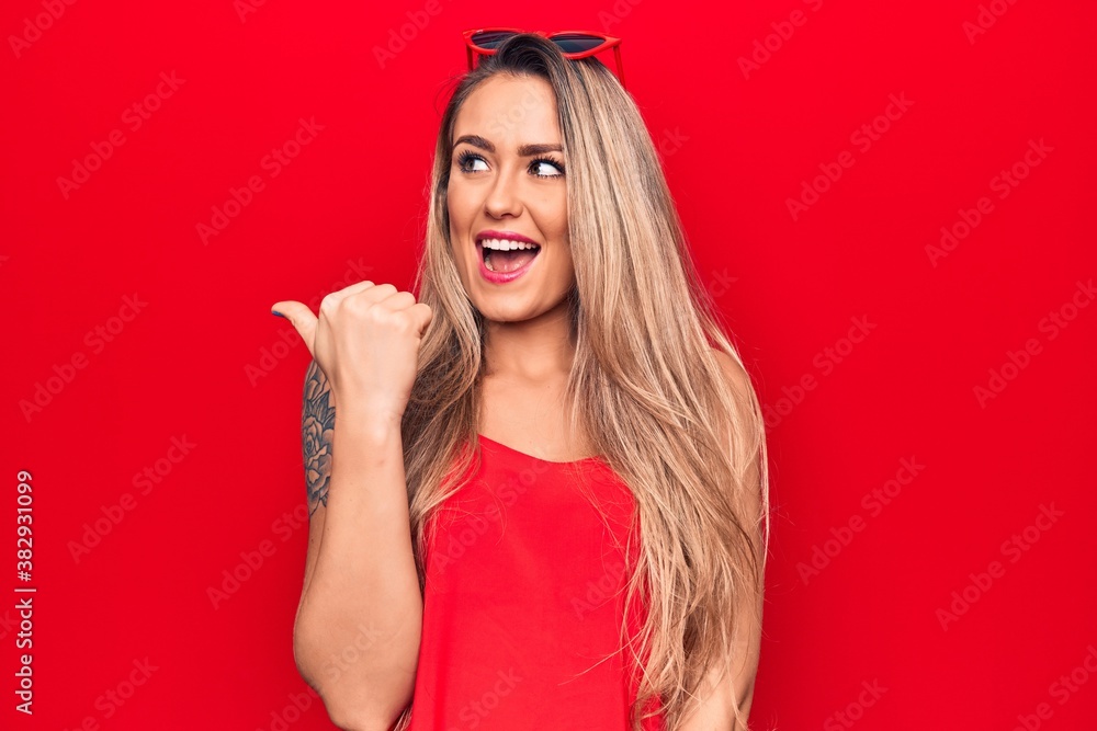 Young beautiful blonde woman wearing red t-shirt and sunglasses over isolated background pointing thumb up to the side smiling happy with open mouth Young beautiful blonde woman wearing red t-shirt and sunglasses over isolated background pointing thumb up to the side smiling happy with open mouth