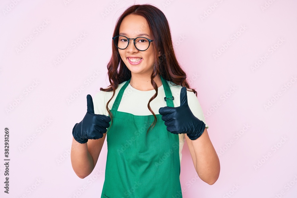Young beautiful chinese girl wearing cleaner apron success sign doing positive gesture with hand, thumbs up smiling and happy. cheerful expression and winner gesture. Young beautiful chinese girl wearing cleaner apron success sign doing positive gesture with hand, thumbs up smiling and happy. cheerful expression and winner gesture.