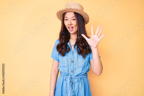 Young beautiful chinese girl wearing summer hat showing and pointing up with fingers number five while smiling confident and happy.