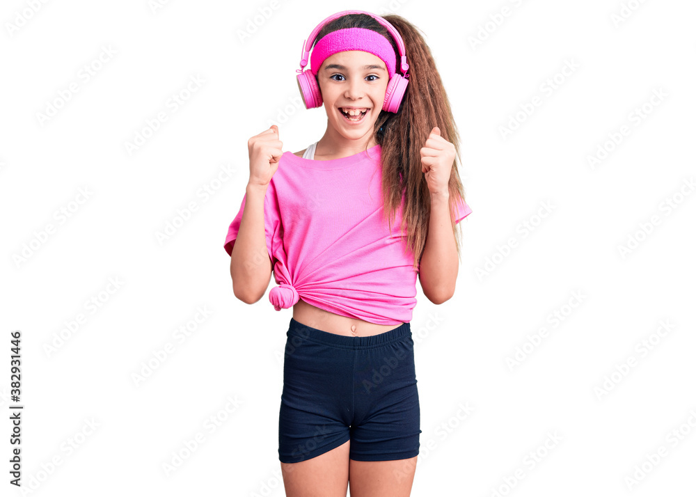 Cute hispanic child girl wearing gym clothes and using headphones celebrating surprised and amazed for success with arms raised and open eyes. winner concept. Cute hispanic child girl wearing gym clothes and using headphones celebrating surprised and amazed for success with arms raised and open eyes. winner concept.