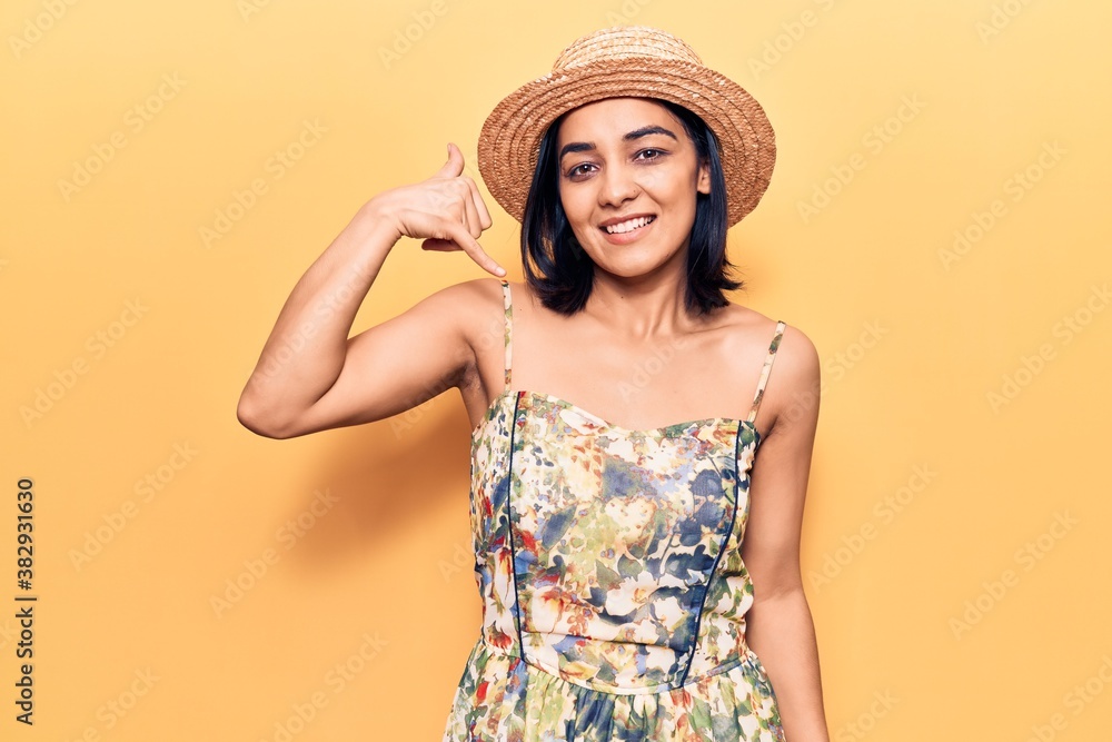 Young beautiful latin woman wearing summer hat smiling doing phone gesture with hand and fingers like talking on the telephone. communicating concepts. Young beautiful latin woman wearing summer hat smiling doing phone gesture with hand and fingers like talking on the telephone. communicating concepts.