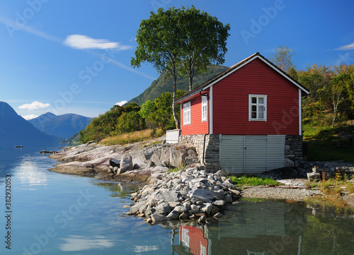 Red Wooden Boat House On The Shore Of The Lustrafjord In Hoyheimsvik On A Sunny Summer Day With A Clear Blue Sky And A Few Clouds