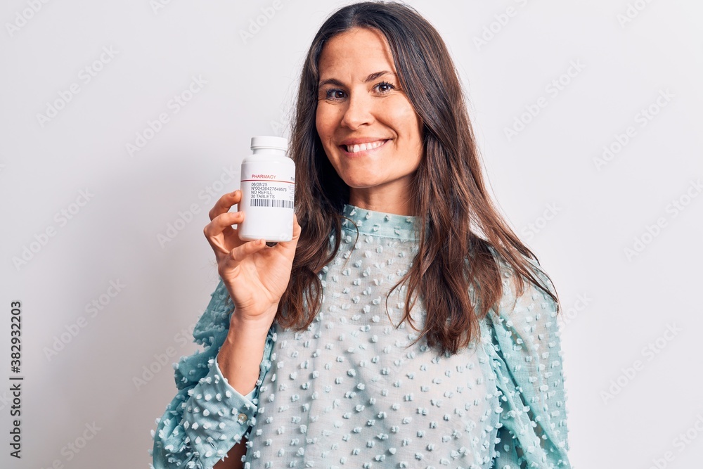 Young beautiful brunette woman holding jar of medicine pills over isolated white background looking positive and happy standing and smiling with a confident smile showing teeth Young beautiful brunette woman holding jar of medicine pills over isolated white background looking positive and happy standing and smiling with a confident smile showing teeth