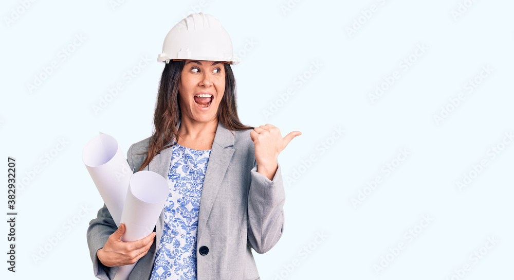 Young beautiful brunette woman wearing architect hardhat holding blueprint pointing thumb up to the side smiling happy with open mouth Young beautiful brunette woman wearing architect hardhat holding blueprint pointing thumb up to the side smiling happy with open mouth