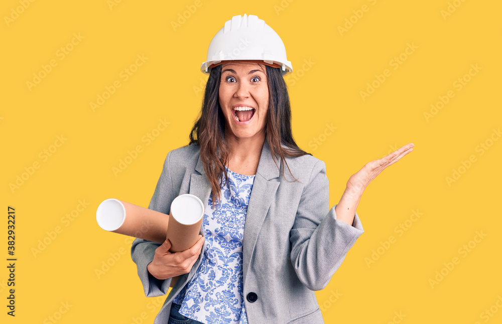 Young beautiful brunette woman wearing architect hardhat holding blueprint celebrating victory with happy smile and winner expression with raised hands Young beautiful brunette woman wearing architect hardhat holding blueprint celebrating victory with happy smile and winner expression with raised hands