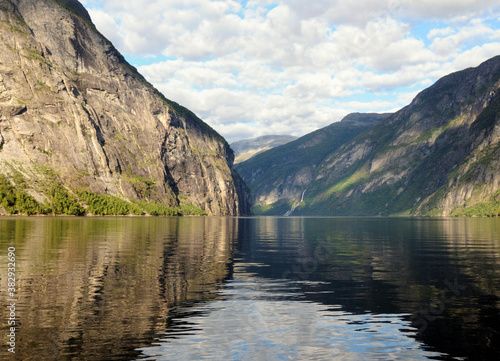 Mountains Reflecting In The Calm Water Of Geirangerfjord On A Sunny Summer Day With A Clear Blue Sky And A Few Clouds