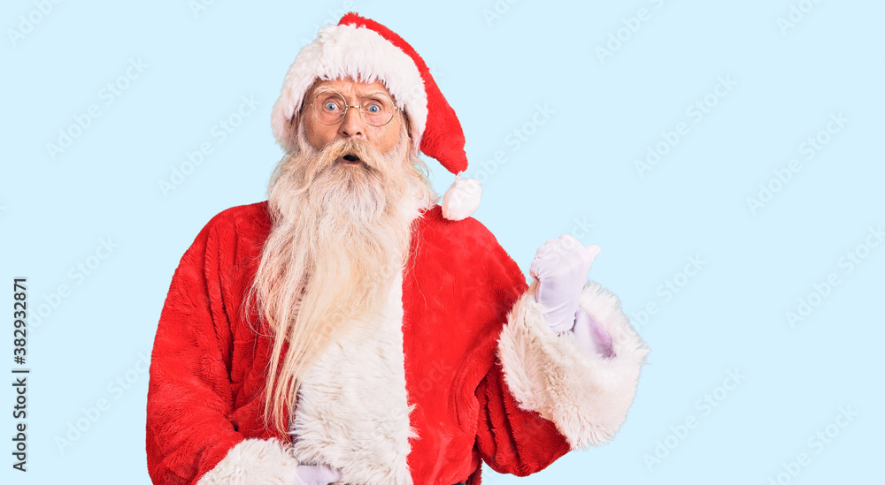 Old senior man with grey hair and long beard wearing traditional santa claus costume surprised pointing with hand finger to the side, open mouth amazed expression. Old senior man with grey hair and long beard wearing traditional santa claus costume surprised pointing with hand finger to the side, open mouth amazed expression.