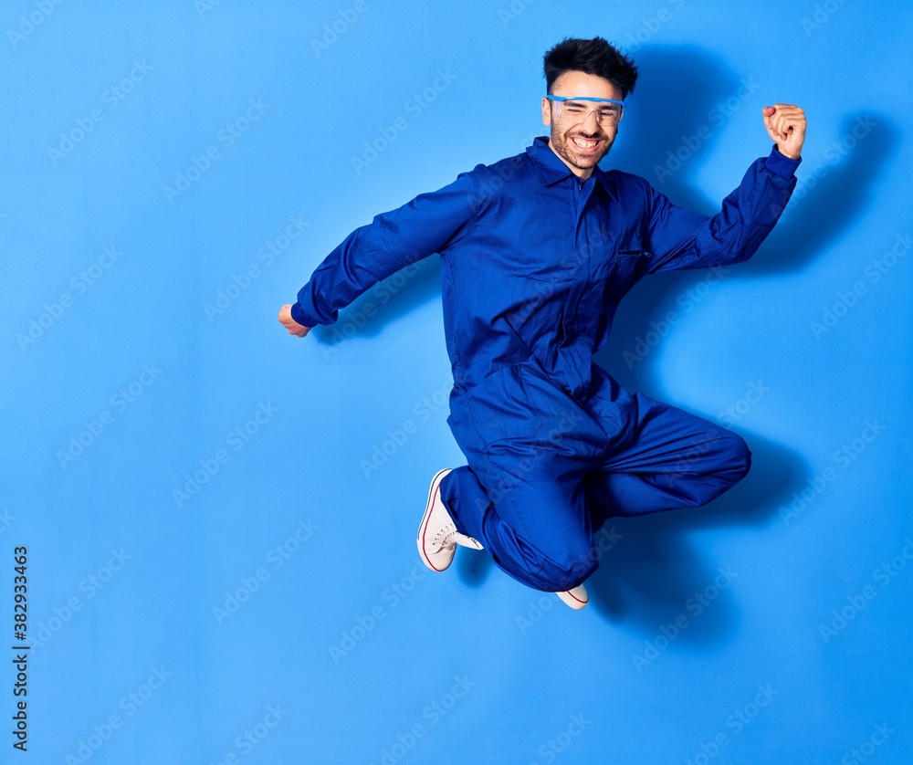 Obraz premium Young handsome hispanic man wearing painter uniform and glasses smiling happy. Jumping with smile on face doig winner sign with fists up over isolated blue background