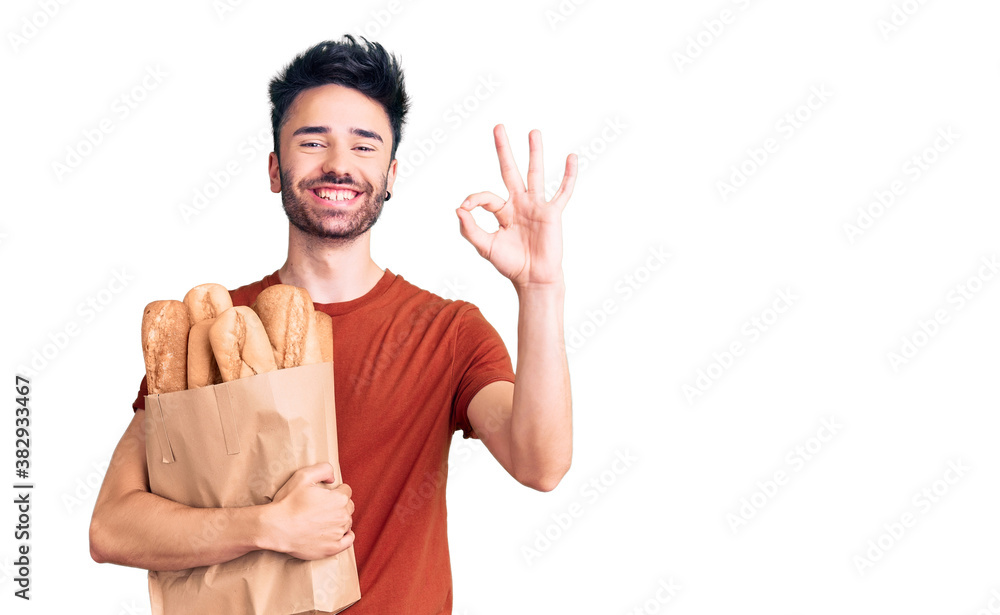 Young hispanic man holding paper bag with bread doing ok sign with fingers, smiling friendly gesturing excellent symbol Young hispanic man holding paper bag with bread doing ok sign with fingers, smiling friendly gesturing excellent symbol