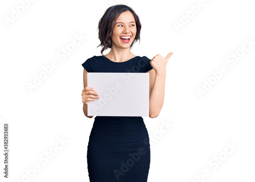 Young beautiful girl holding blank empty banner pointing thumb up to the side smiling happy with open mouth