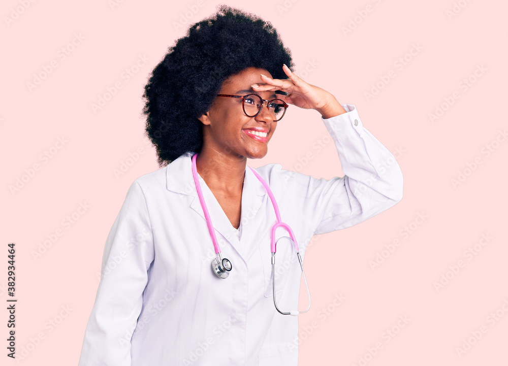 Young african american woman wearing doctor coat and stethoscope very happy and smiling looking far away with hand over head. searching concept.