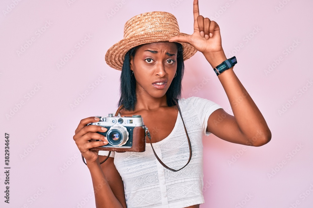 Young african american woman wearing summer hat holding vintage camera making fun of people with fingers on forehead doing loser gesture mocking and insulting.