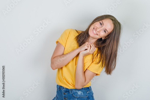 Young woman posing like asking for a favor in yellow t-shirt, shorts and looking cheerful , front view.